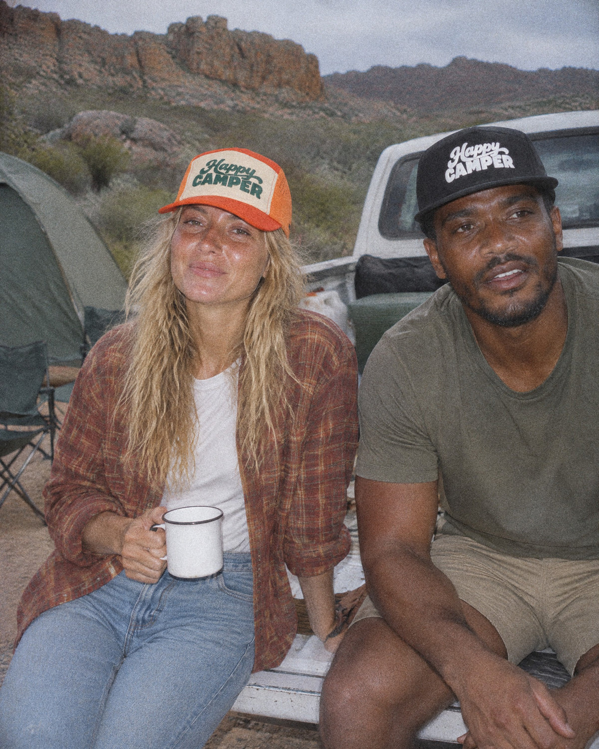Two friends at a campsite in black and orange Happy Camper caps